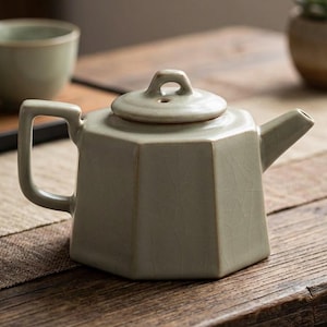 Puede incluir: Una tetera de cerámica verde claro con un diseño geométrico. La tetera tiene una tapa con un asa pequeña y un pico. El asa es rectangular. La tetera está sobre una superficie de madera. Una taza pequeña está en el fondo.