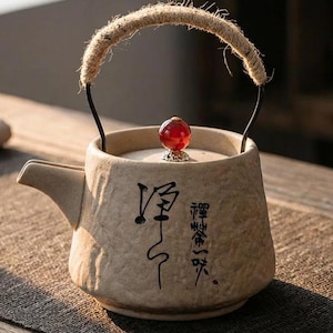 May include: A beige ceramic teapot with a rope handle and a red gem on the lid. The teapot has black calligraphy on the front. The spout is on the left side. The handle is made of a thick rope.