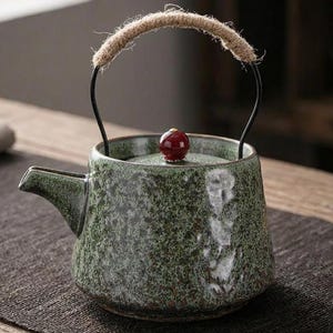 以下が含まれることがあります： テクスチャ加工された表面とロープのハンドルが付いた緑色のセラミックティーポット。ティーポットには小さな注ぎ口と蓋に赤いノブが付いています。ハンドルは、ロープが巻き付けられた黒い金属フレームで作られています。