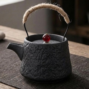 Puede incluir: Tetera de cerámica negra con superficie texturizada y boquilla. El asa es de metal negro y está envuelta con cuerda natural. Un botón redondo rojo adorna la tapa. Diseñada para preparar té.