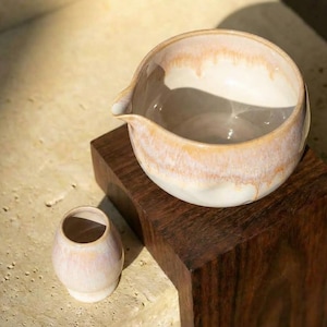 May include: A ceramic tea set featuring a large, round pitcher with a spout and a small, cylindrical cup. The pitcher and cup have a white base with a peach and tan glaze. The set is displayed on a dark wooden block.