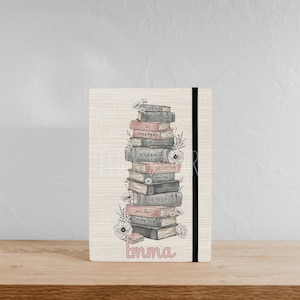 May include: A beige journal featuring a stack of books illustration. The books are in shades of grey, pink, and brown, with floral accents. The text on the books includes phrases like "Life is like a book" and "Emma". A black elastic band is on the right side.