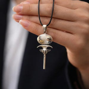 Puede incluir: Collar con colgante de plata con cordón negro. El colgante presenta una parte superior ovalada y un diseño estilizado debajo. El collar se muestra sobre un fondo oscuro.