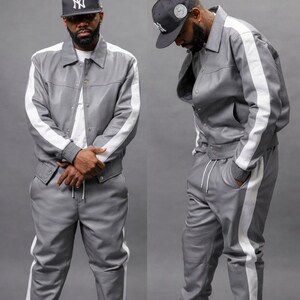 Puede incluir: Un ch&aacute;ndal de cuero gris con rayas blancas en los lados de la chaqueta y los pantalones. La chaqueta tiene cierre de botones y los pantalones tienen cintura con cord&oacute;n. Se lleva una gorra de b&eacute;isbol negra con un logotipo blanco.