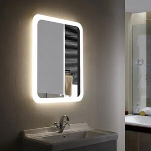 Peut inclure: Un miroir de salle de bain rectangulaire éclairé avec des coins arrondis. Le miroir a une bordure LED blanche et lumineuse et est monté au-dessus d'un lavabo blanc avec un robinet chromé. Une serviette est suspendue à un porte-serviettes derrière le miroir.