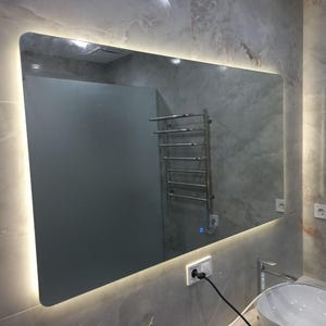 May include: A rectangular bathroom mirror with rounded corners and a built-in LED light. The mirror is mounted on a gray tiled wall, reflecting a towel rack and a sink. The overall design is modern and minimalist.