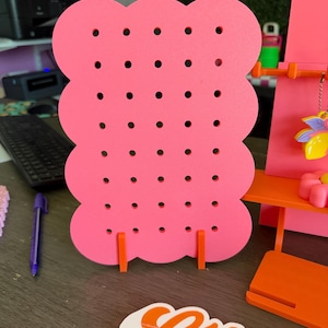 May include: A pink earring display stand with a cloud-like shape and multiple holes for hanging earrings. The stand is supported by two orange legs. Other accessories are visible in the background.
