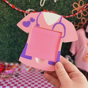 May include: A pink nurse-themed photo frame with purple accents, including a stethoscope and heart shapes. The frame has a rectangular opening for a photo and is held by a hand. The background includes the text "The Lily Barn".