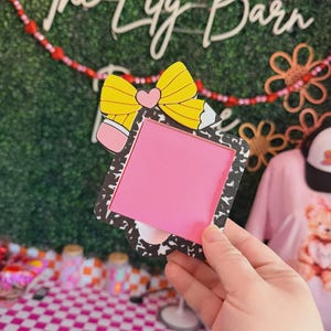 May include: A pink square notepad with a yellow pencil bow design. The notepad is held in front of a green background with the words "The Lily Barn" in white script. A pink and white checkered table is in the foreground.