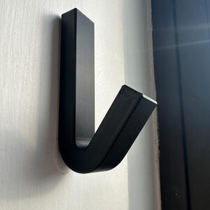 Op de afbeelding: Een zwarte, moderne wandhaak aan een witte muur. De haak heeft een rechthoekig profiel met een gebogen onderkant en een rechte bovenkant, waardoor een J-vorm ontstaat. De haak is gemaakt van een solide, matzwart materiaal.
