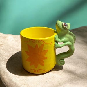 May include: A bright yellow ceramic mug featuring a hand-painted orange sun design on the front. The handle is sculpted into the shape of a light green chameleon with detailed eyes, clinging to the side of the mug with its claws and curled tail.