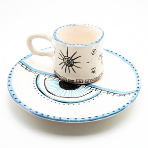 May include: A white ceramic cup and saucer set featuring hand-painted black and blue designs. The cup displays a sun motif and small symbols, while the saucer features a large stylized eye graphic with a blue iris and a dotted border pattern.