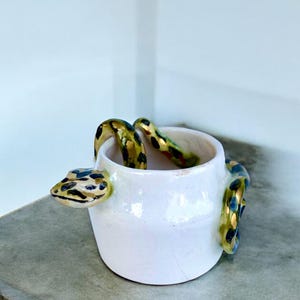 Puede incluir: Una pequeña taza de cerámica blanca con una serpiente esculpida envuelta alrededor del exterior. La serpiente tiene un acabado brillante con un patrón de manchas negras y doradas. La taza tiene un cuerpo liso y redondeado con una abertura ancha.