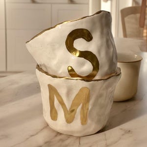 May include: Two stacked white ceramic bowls with an uneven, textured surface and gold-painted rims. The top bowl features a large gold letter S, while the bottom bowl displays a large gold letter M. Both bowls have a glossy finish and organic shape.
