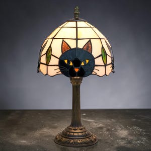 May include: A Tiffany-style table lamp featuring a black cat design on the shade. The shade is composed of stained glass in cream, black, brown, and green. The base is bronze-colored with an ornate, floral design.