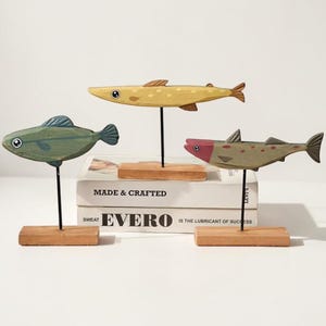 Peut inclure: Trois sculptures de poissons en bois, vertes, jaunes et marron, chacune montée sur une base en bois avec une tige métallique noire. Les poissons sont exposés sur une surface blanche, avec des livres en arrière-plan avec les mots "MADE & CRAFTED" et "EVERO".