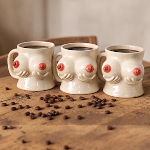May include: Three cream-colored ceramic mugs shaped like breasts, each with a red nipple. The mugs are filled with dark coffee and have handles. Coffee beans are scattered on the wooden surface.