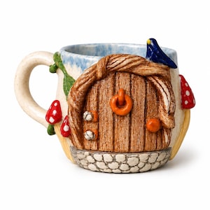 Può includere: Una tazza in ceramica con una porta in legno marrone scolpita, una maniglia arancione, una base in pietra e funghi rossi con macchie bianche. La tazza presenta un interno azzurro e bianco, un manico curvo e un piccolo uccellino blu sopra.