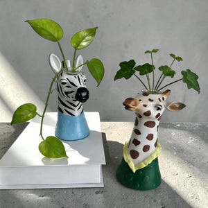 Könnte beinhalten: Zwei dekorative Vasen in Tierform, eine Zebra- und eine Giraffenvase, die jeweils grüne Pflanzen enthalten. Die Zebra-Vase hat einen schwarz-weiß gestreiften Kopf und einen blauen Sockel. Die Giraffenvase hat braune Flecken und einen grünen Sockel.