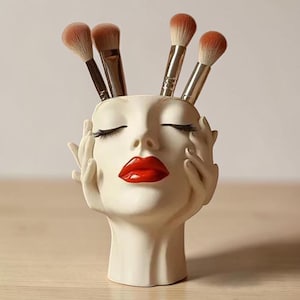 May include: A cream-coloured face-shaped makeup brush holder with red lips and closed eyes, holding several makeup brushes. The holder has hands on each side of the face. The brushes have brown bristles and silver handles.