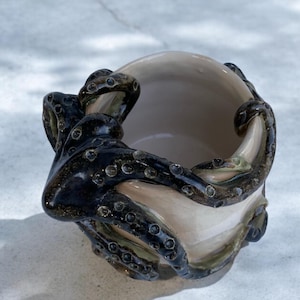May include: A ceramic mug with a smooth, light beige interior and exterior. The exterior is wrapped in a dark, glossy, black-glazed octopus sculpture with textured suckers that extend around the sides and rim of the vessel.