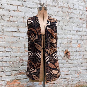May include: A long, open-front vest with a brown, black, and beige geometric pattern. The vest is displayed on a vintage dress form with a metal stand, set against a weathered brick wall.