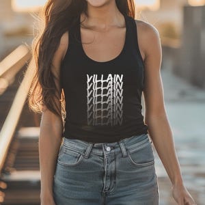 May include: Black vest top featuring the word "VILLAIN" in a stacked, white and grey gradient design. The top is worn with blue denim jeans. The image is well-lit, highlighting the casual style.