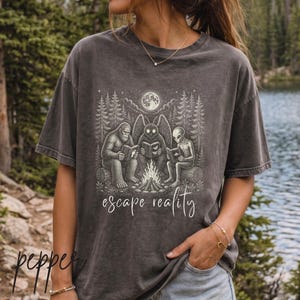 Op de afbeelding: Een antracietgrijs t-shirt met korte mouwen en een witte grafische print van Bigfoot, een mothman en een alien die in een bos onder een volle maan rond een kampvuur zitten te lezen, met de tekst escape reality onder de afbeelding.