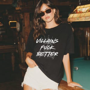 May include: Black t-shirt with the text "Villains Fuck Better" in white, featuring a skull and crossbones graphic. The shirt is worn with white shorts. The image is taken in a dimly lit room with a pool table in the background.