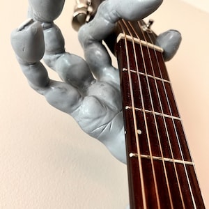 May include: A gray, hand-shaped guitar holder with a dark brown fretboard and strings. The holder is mounted on a wall, showcasing the guitar's neck and headstock. The design is artistic and unique, perfect for displaying a guitar.