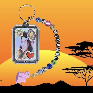 May include: A silver keychain with a rectangular pendant featuring a photo of Michael Jackson, surrounded by decorative stickers. The keychain includes a beaded strand spelling "MICHAEL" and a pink animal charm. The background is an orange sunset.