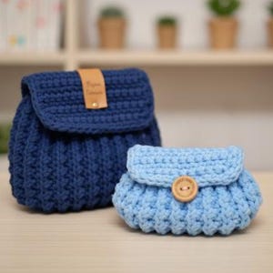 Pode incluir: Duas bolsas de crochê feitas à mão. Uma bolsa maior azul marinho estilo mochila com fecho de aba e uma pequena etiqueta de couro marrom. Uma embreagem azul claro menor com fecho de botão.
