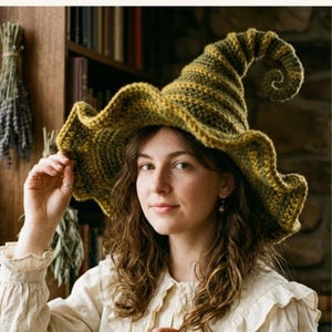 May include: A handmade, olive green and brown crochet witch hat with a wavy brim and a curled tip. The hat is worn by a person with long, wavy brown hair. The background includes a bookshelf and dried herbs.