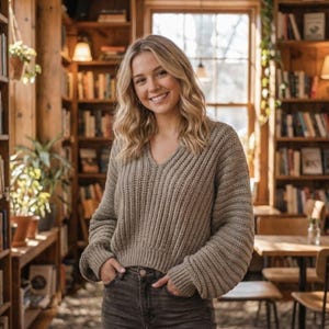 May include: A woman wearing a tan, ribbed knit sweater with a V-neck and long sleeves, standing in a library. She is smiling and has her hands in her pockets. The background includes bookshelves, plants, and a window.