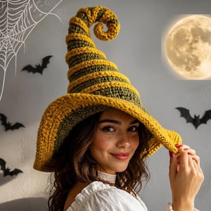 May include: A crocheted witch hat featuring a tall, spiralled point and a wide brim. The hat is constructed with alternating horizontal stripes of mustard yellow and olive green yarn, creating a textured, ribbed appearance throughout the design.