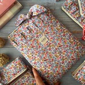 Könnte beinhalten: Laptop-Hülle mit Blumenmuster und passendes Zubehör. Die Hülle und das Zubehör weisen ein Muster aus kleinen roten, gelben und blauen Blumen auf hellblauem Hintergrund auf. Das Zubehör umfasst eine Reißverschlusstasche und eine Schleife.