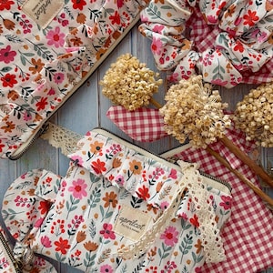 Op de afbeelding: Cosmetische tassen, etuis en scrunchies met bloemenprint. De items hebben een witte achtergrond met rode, roze en oranje bloemen. De tassen hebben een ritssluiting en een kanten rand, met een ruche detail.
