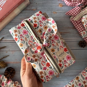 Könnte beinhalten: Eine Reißverschlusstasche mit floralem Druck und Schleifendetail. Die Tasche hat einen cremefarbenen Hintergrund mit roten, rosa und orangefarbenen Blumen. Der Reißverschluss ist hellbraun. Die Tasche ist mit "apricot" beschriftet.