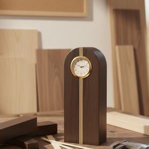 Könnte beinhalten: Eine dunkelbraune Holzuhr mit goldfarbenem Zifferblatt. Die Uhr hat einen vertikalen Goldstreifen auf jeder Seite. Die Uhr steht auf einer Holzwerkbank mit Werkzeugen und Holzstücken.