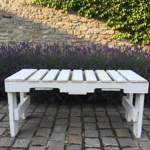 Op de afbeelding: Een witte houten bank met een lattenbodem en stevige poten. De bank is wit geschilderd en staat op een geplaveide ondergrond, met een achtergrond van een stenen muur, paarse bloemen en groen gebladerte.