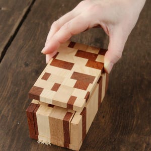 Può includere: Una scatola di puzzle in legno con un design geometrico. La scatola è realizzata con pezzi di legno marrone chiaro e scuro, con un coperchio che viene sollevato. La scatola è su una superficie di legno marrone scuro.