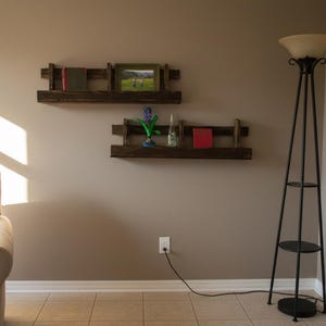 May include: Two rustic wooden floating shelves mounted on a neutral-colored wall. The top shelf holds a framed picture and a book, while the bottom shelf displays a small vase, a book, and a decorative item. A black floor lamp stands nearby.