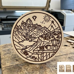 Könnte beinhalten: Eine runde Holzszene mit Campingmotiv, die eine Bergkette, Zelte und eine Kaffeekanne zeigt. Das Design ist in das Holz geätzt, mit einer Sonne und einem Gleitschirm im Himmel. Das Holz ist hellbraun und liegt auf einem Holztisch.