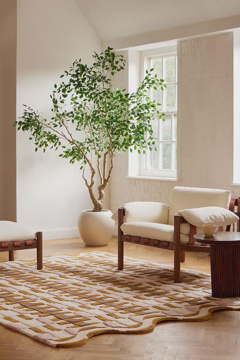 May include: An interior scene featuring a large potted tree with green leaves, a cream-colored armchair with brown leather straps, and a patterned rug in shades of beige, pink, and brown. A small side table and a window are also visible.