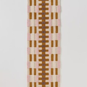 May include: A rectangular rug with a woven pattern in shades of tan, brown, pink, and cream. The rug has a wavy top and bottom edge. The design features vertical and horizontal blocks of color, creating a textured surface.