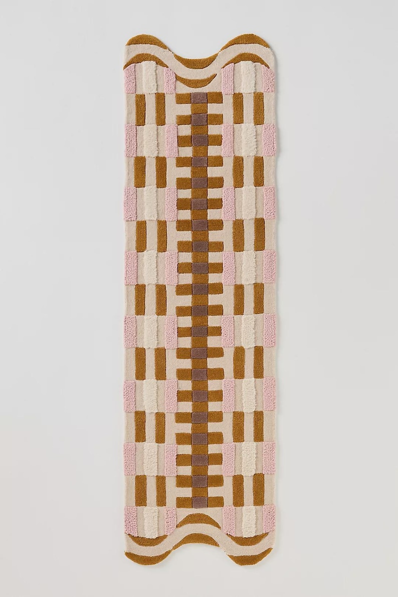 May include: A rectangular rug with a woven pattern in shades of tan, brown, pink, and cream. The rug has a wavy top and bottom edge. The design features vertical and horizontal blocks of color, creating a textured surface.