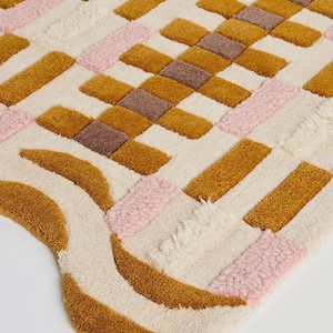 May include: A colorful, textured rug with a geometric pattern. The rug features a cream-colored background with sections of mustard yellow, pink, and brown rectangles and curved shapes. The rug is made of a soft, plush material.