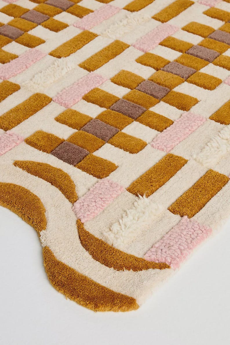 May include: A colorful, textured rug with a geometric pattern. The rug features a cream-colored background with sections of mustard yellow, pink, and brown rectangles and curved shapes. The rug is made of a soft, plush material.
