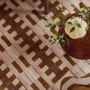 May include: A geometric patterned rug in shades of brown, pink, and cream. The rug features a woven design with rectangular and square shapes. A wooden chair leg and a small table with a vase of flowers are visible in the background.
