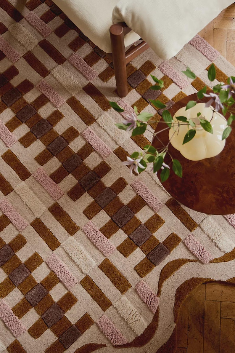 May include: A geometric patterned rug in shades of brown, pink, and cream. The rug features a woven design with rectangular and square shapes. A wooden chair leg and a small table with a vase of flowers are visible in the background.